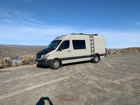Explore with this beige Mercedes Sprinter RV rental. Parked on a gravel lot overlooking an arid landscape and river.