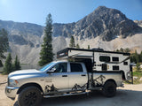 Silver Ram truck with a Palomino truck camper setup by a stunning mountain lake, ideal for an RV rental adventure.