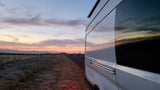 A white RV rental on a roadside, its windows reflecting a vivid sunset over mountains and a desert landscape.