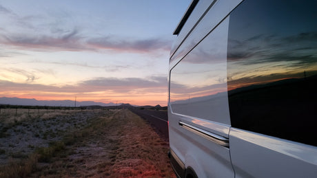 A white RV rental on a roadside, its windows reflecting a vivid sunset over mountains and a desert landscape.
