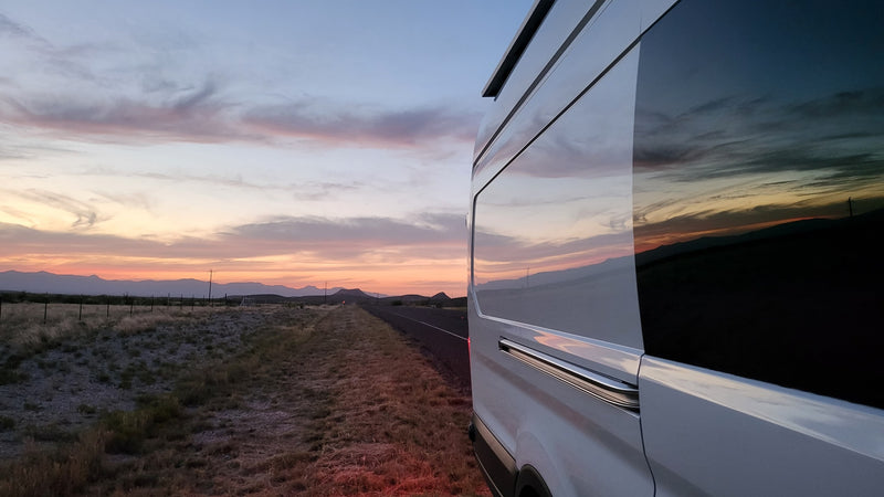 A white RV rental on a roadside, its windows reflecting a vivid sunset over mountains and a desert landscape. — photo 9