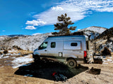 RV rental: Silver Winnebago Revel 4x4 camper van parked on a snowy mountain road under a blue sky, with camp chairs nearby.