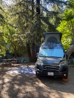 This dark camper van RV rental is perfectly parked in a sunny, wooded campsite, ready for outdoor adventure.