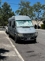 Grey Mercedes-Benz Sprinter camper van, possibly an RV rental, with a sunshade parked on a sunny street.