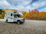 Scenic RV rental parked by a road, surrounded by vibrant autumn trees and mountains under a cloudy sky. Fall road trip!