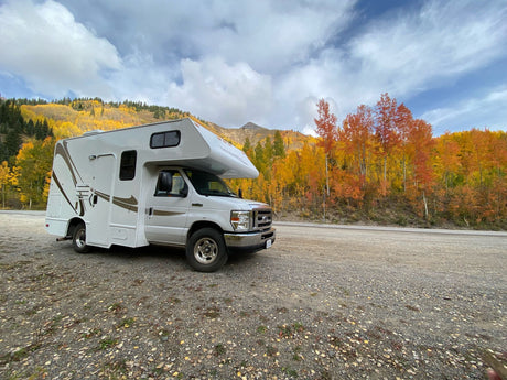 Scenic RV rental parked by a road, surrounded by vibrant autumn trees and mountains under a cloudy sky. Fall road trip!