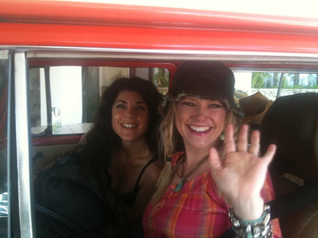 Two smiling women inside an RV rental. The blonde woman in a hat waves, while her dark-haired friend smiles nearby.