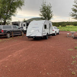This RV rental site features a FREESOLO travel trailer, white SUV, and gray pickup parked on red gravel with trees.