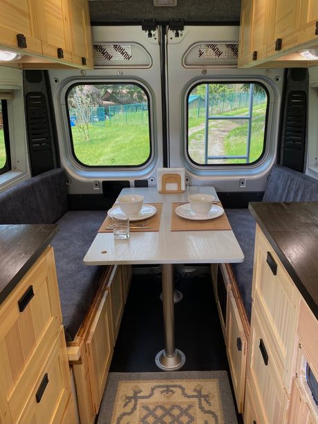 Cozy rv rental dining area in a camper van, featuring a table, gray benches, and light wood cabinetry with outdoor views.