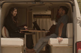 Inside a cozy rv rental, a couple smiles while sitting at a table with laptops in their camper van living space.