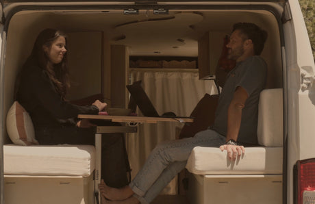 Inside a cozy rv rental, a couple smiles while sitting at a table with laptops in their camper van living space.
