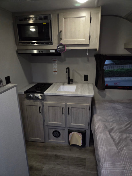 Compact rv rental kitchen: microwave, stove, sink, and cabinets, with a partial view of the bed. Efficient travel living.
