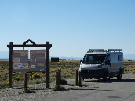An RV rental camper van at a roadside historical sign ("On Sacred Ground") in a vast, sunny, open desert landscape.