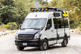 White Mercedes-Benz Sprinter adventure van RV rental, equipped with roof rack, lights, and off-road tires, parked on a road.