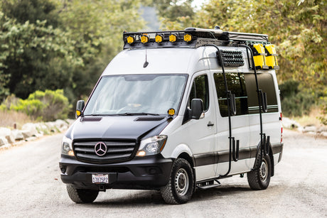 White Mercedes-Benz Sprinter adventure van RV rental, equipped with roof rack, lights, and off-road tires, parked on a road.