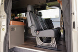 This RV rental camper van interior offers black passenger seats, a compact kitchen with a brown cushion, and wood-look flooring.