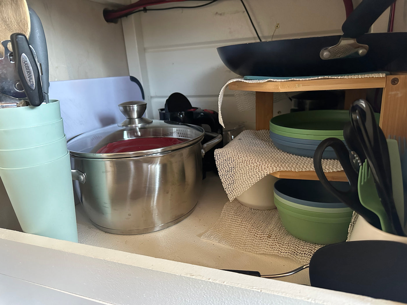 Organized RV rental kitchen: a cabinet stocked with a pot, pan, stacked green/blue dishes, cups, and utensils for compact travel.