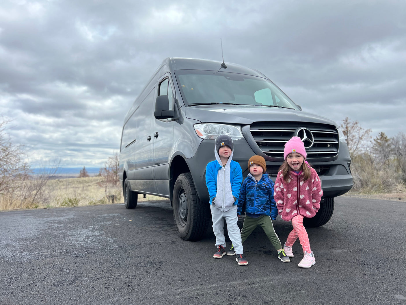 Three joyful kids stand before a grey Mercedes Sprinter RV rental van on a cloudy day, ready for adventure.