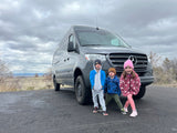 Three joyful kids stand before a grey Mercedes Sprinter RV rental van on a cloudy day, ready for adventure.