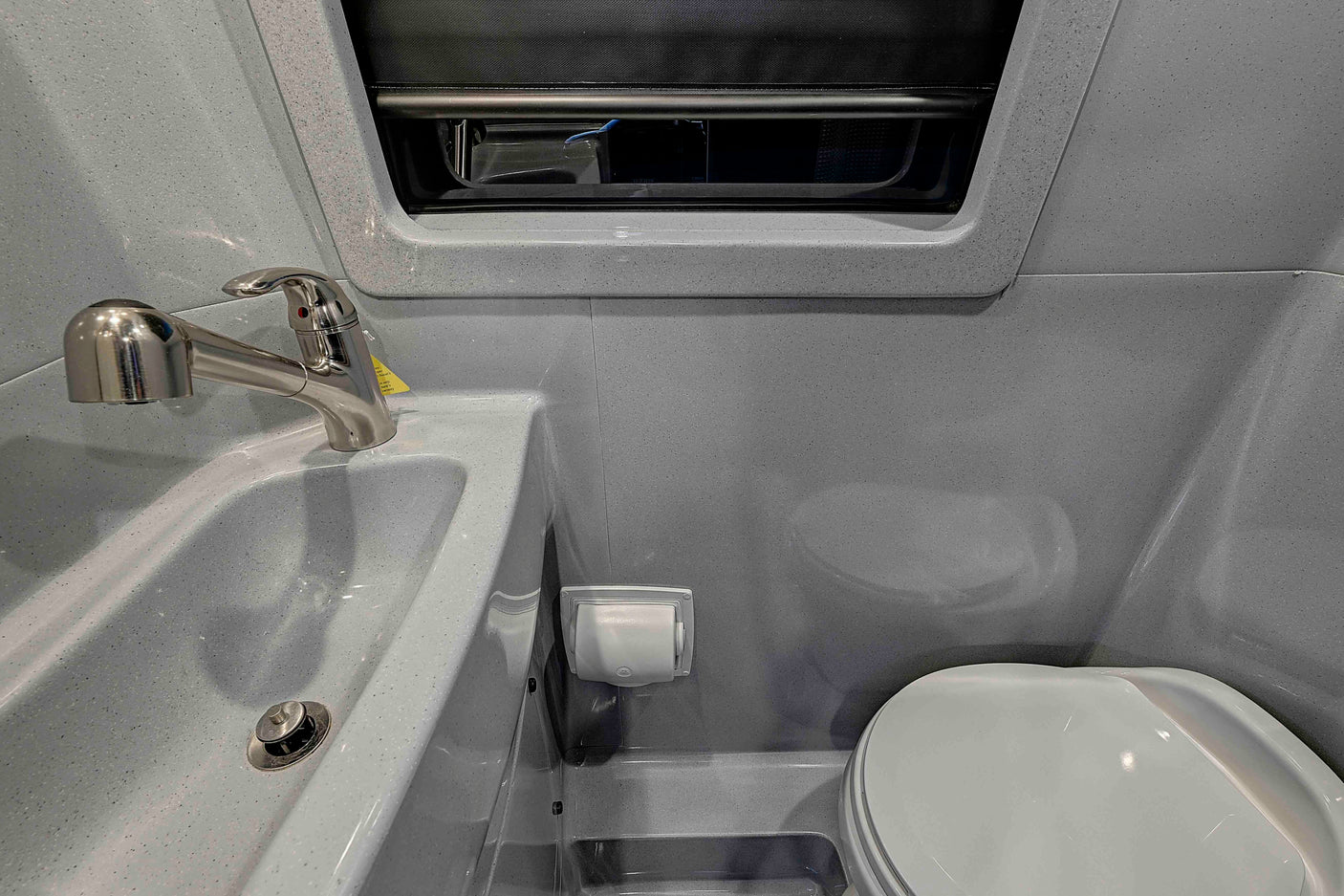 Compact gray speckled RV bathroom for rv rental, featuring a sink with a pull-out faucet, toilet, and window blind.