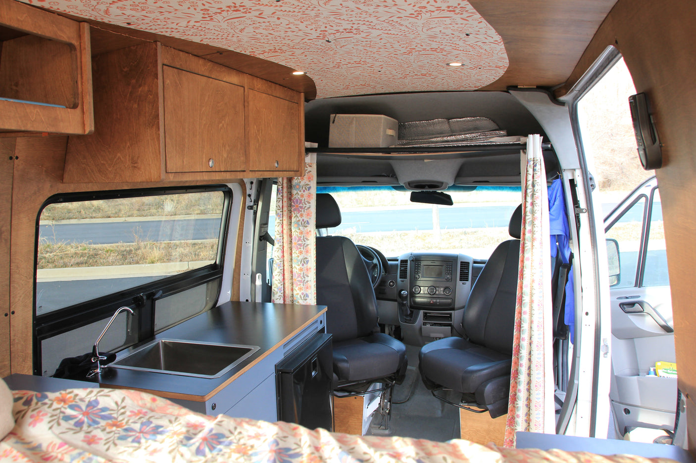 View inside an RV rental showing a wood-paneled kitchen with sink, built-in fridge, floral bed, and front driver's cab.