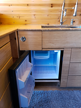 Modern RV rental kitchen featuring a compact fridge with a glowing blue interior, wood cabinetry, sink, and rug.