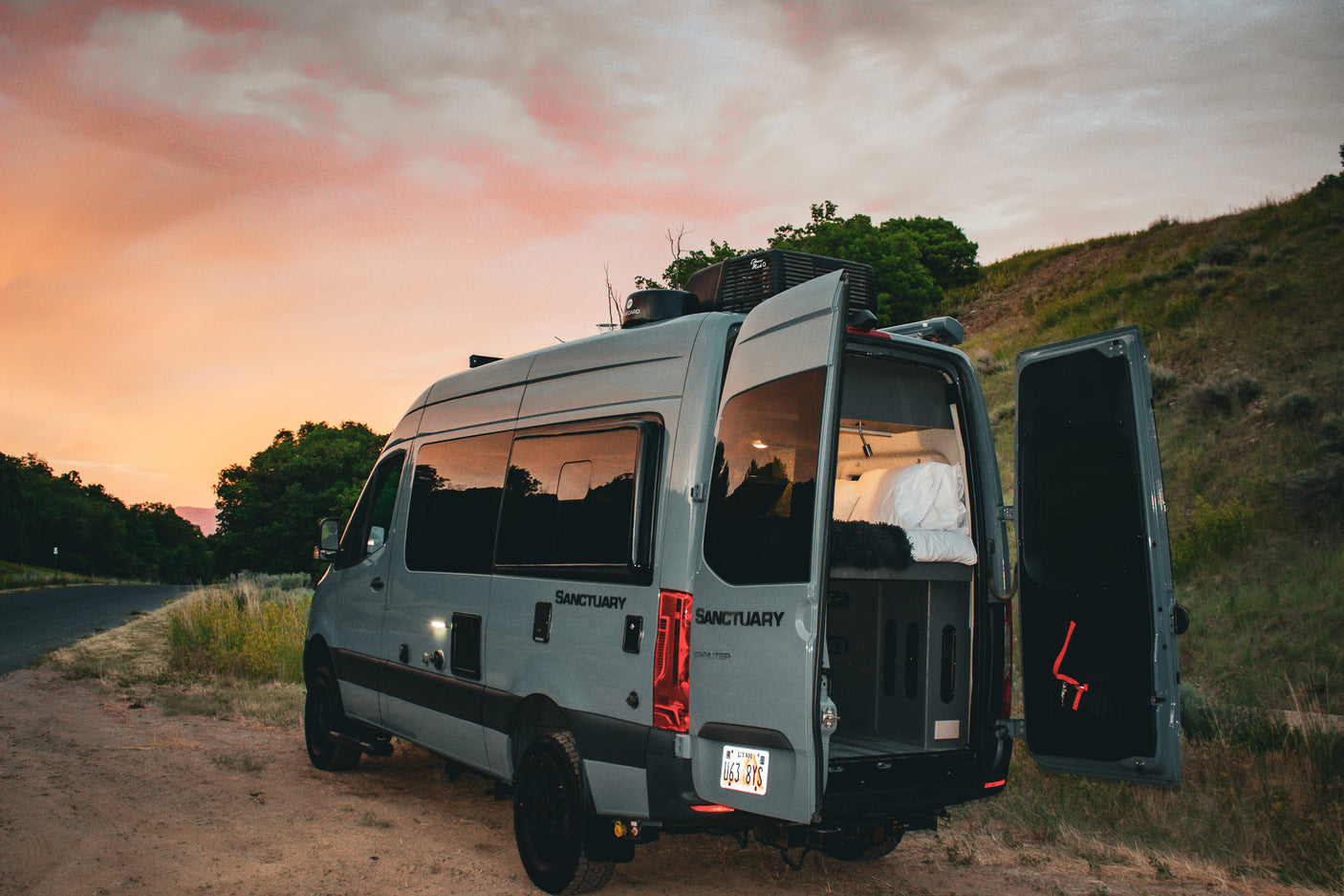 A grey camper van with open rear doors showing a cozy bed, parked at sunset. Ideal for your next RV rental outdoor adventure.