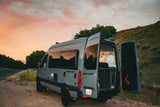 A grey camper van with open rear doors showing a cozy bed, parked at sunset. Ideal for your next RV rental outdoor adventure.