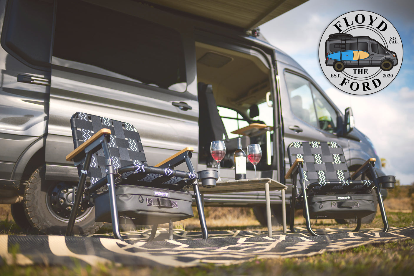 Cozy RV rental setup: a camper van with extended awning, two camping chairs, a table with wine, and an outdoor mat for relaxation.