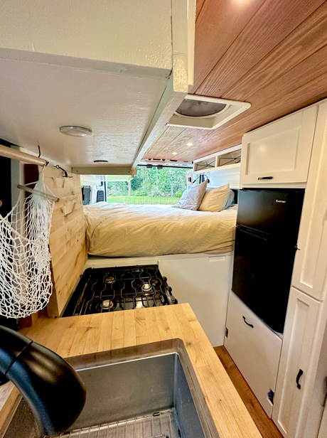 Spacious RV rental interior with a cozy bed at the back, kitchen area featuring a sink & stove, and a mini-fridge.