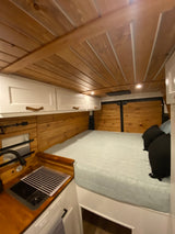 Well-appointed RV rental camper van interior. Shows a comfortable bed, wood-paneled walls/ceiling, white cabinets, and a kitchen sink.