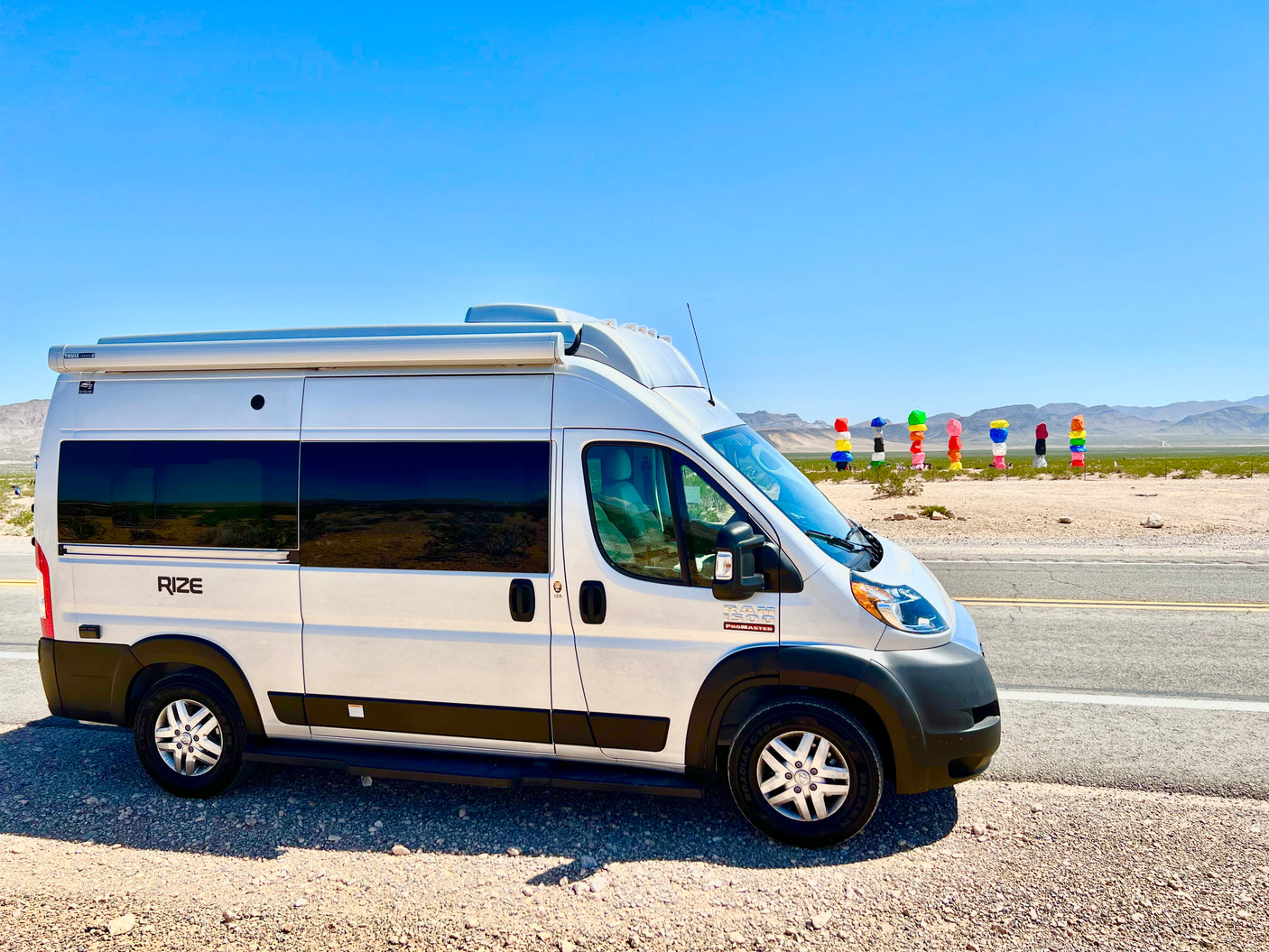 White Ram ProMaster RV rental parked by a desert road with vibrant Seven Magic Mountains art in the background.