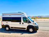 White Ram ProMaster RV rental parked by a desert road with vibrant Seven Magic Mountains art in the background.