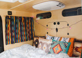 Cozy RV rental with a fluffy white bed, colorful Southwestern pillows, and purple string lights in a wood-paneled interior.