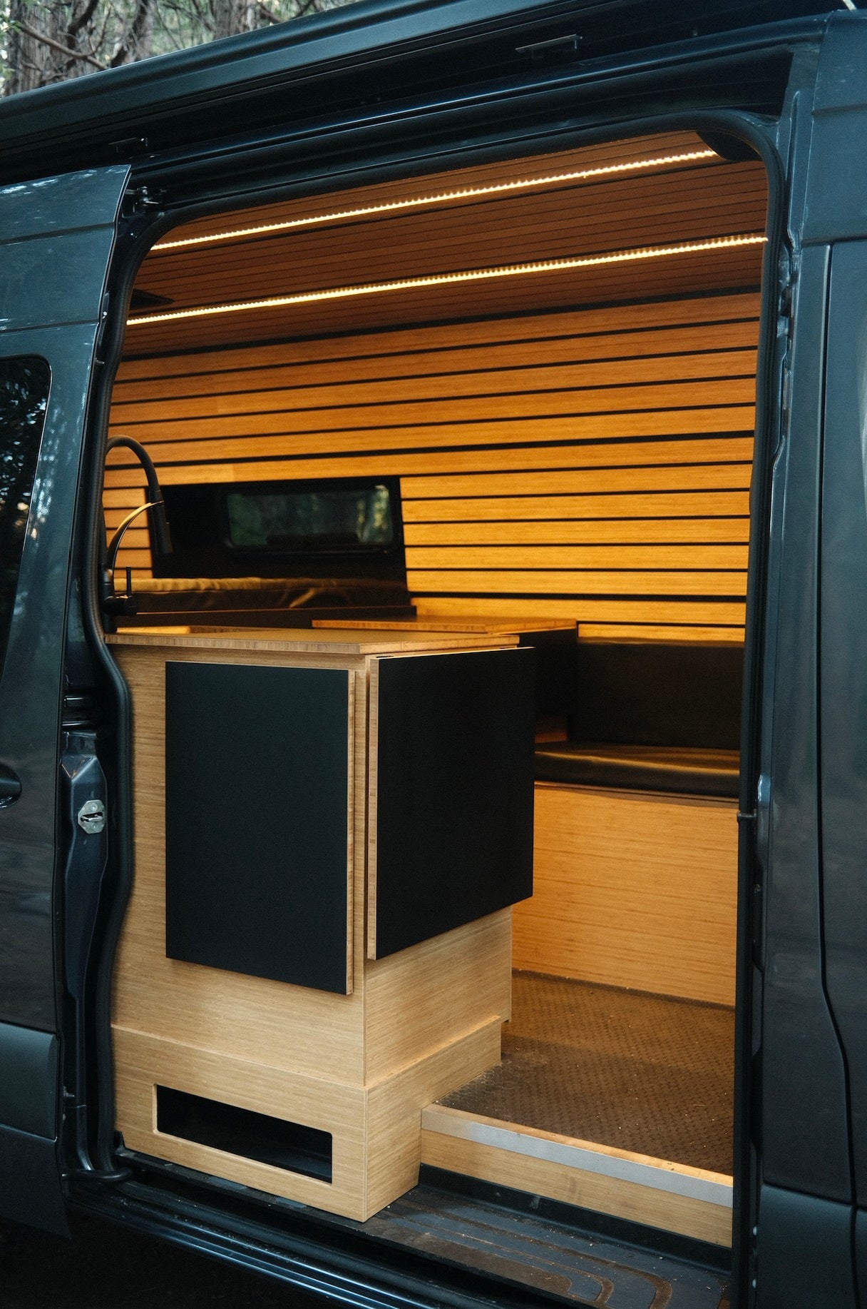 Modern rv rental camper van interior: light wood walls, LED lighting, compact kitchen with black cabinets, and bench.