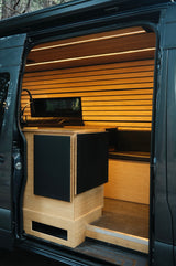 Modern rv rental camper van interior: light wood walls, LED lighting, compact kitchen with black cabinets, and bench.