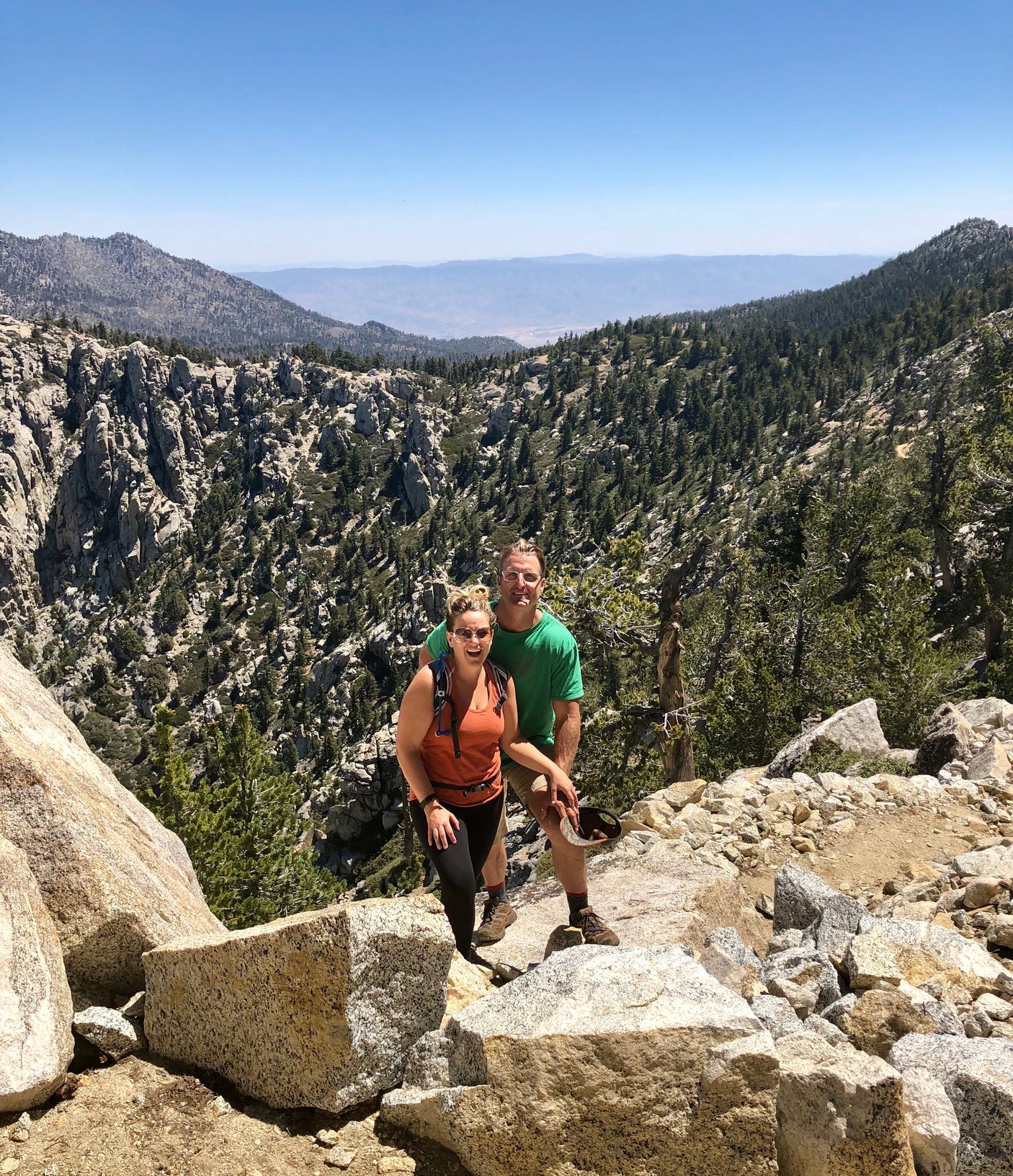 A couple smiles on a sunny mountain hike, overlooking a vast, treed valley. Ideal for an **rv rental** outdoor adventure.