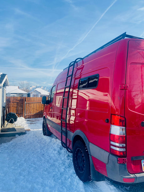 Red Sprinter RV rental van, with roof rack and ladder, parked in a snowy winter yard with mountains in background.