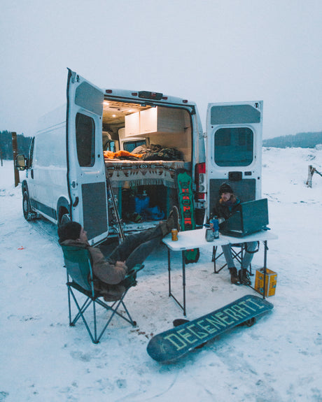 Cozy RV rental experience: Two people, one on a laptop, relax outside their camper van in a snowy landscape. Snowboards nearby.