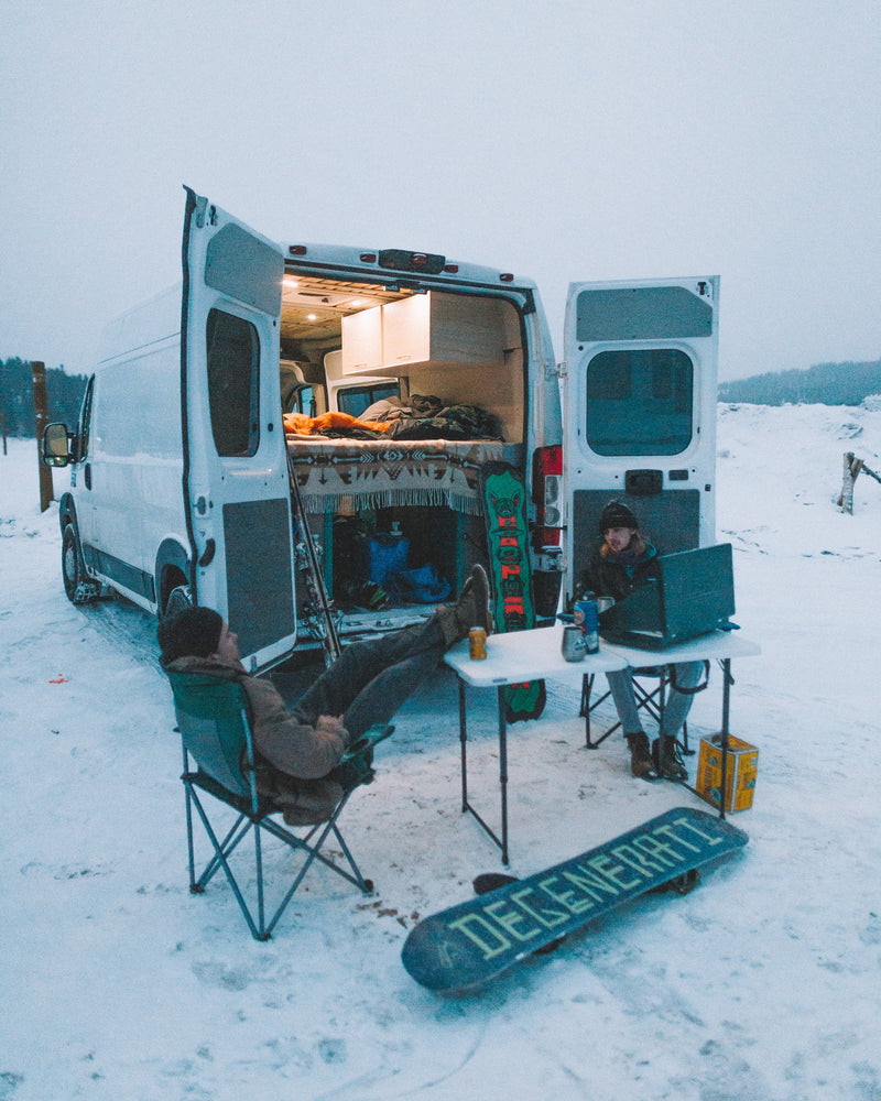 Cozy RV rental experience: Two people, one on a laptop, relax outside their camper van in a snowy landscape. Snowboards nearby. — photo 13