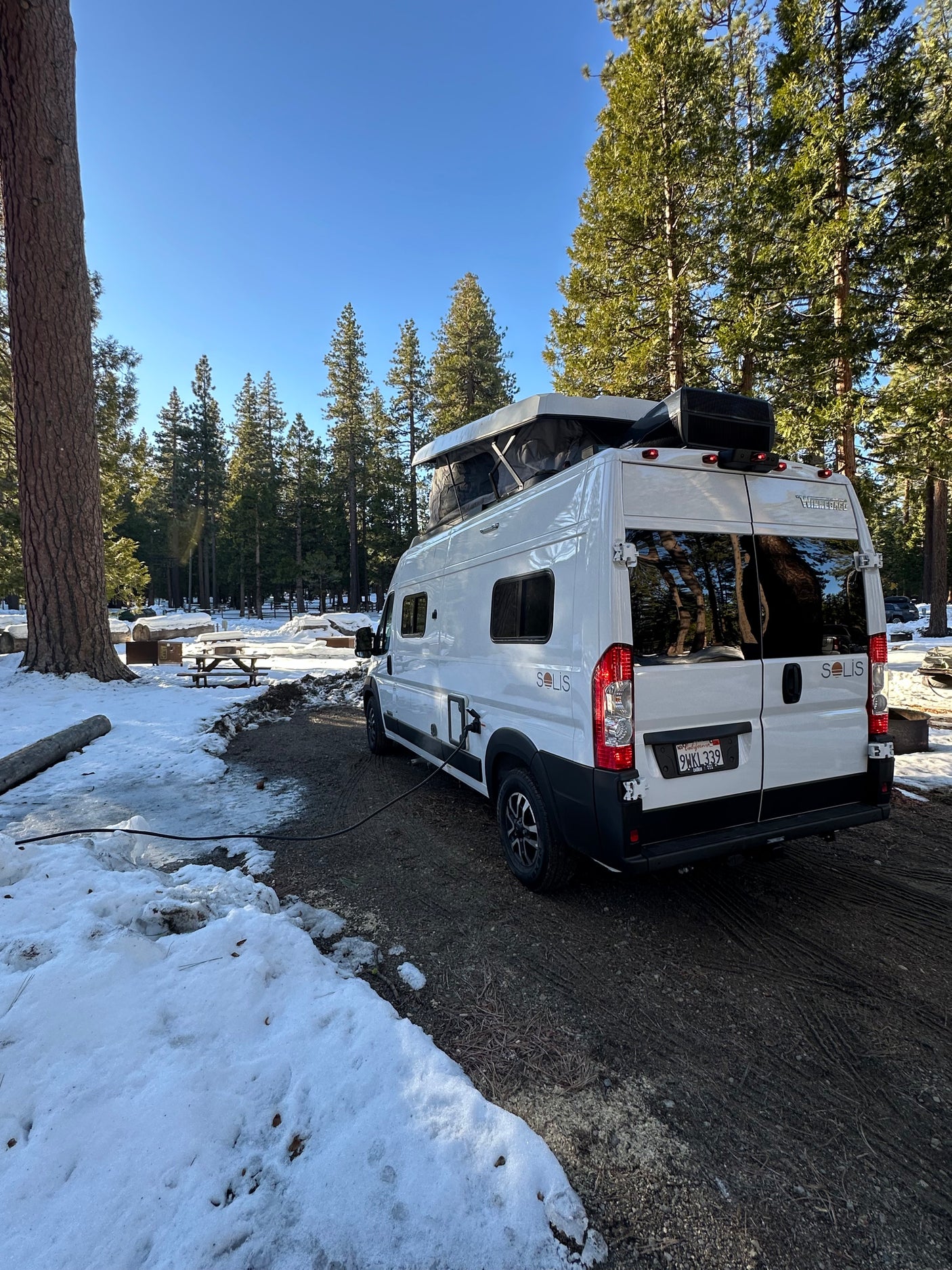 White Winnebago Solis rv rental parked in a snowy forest campground with pop-top open, power cord connected for winter camping.