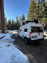 White Winnebago Solis rv rental parked in a snowy forest campground with pop-top open, power cord connected for winter camping.