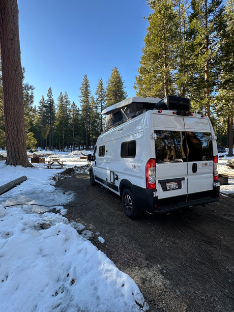 White Winnebago Solis rv rental parked in a snowy forest campground with pop-top open, power cord connected for winter camping.