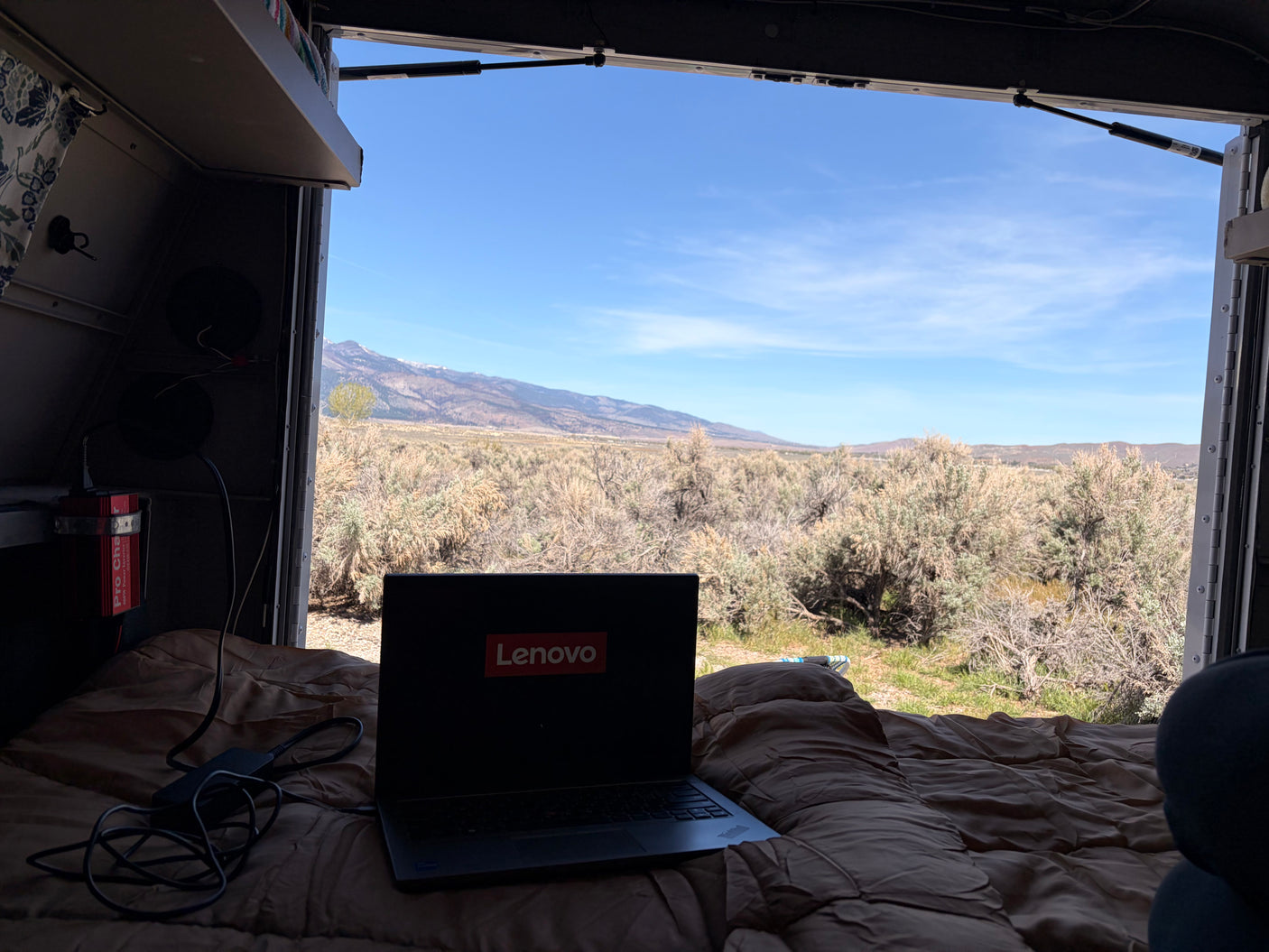 Working remotely from an RV rental, enjoying desert mountains through the open hatch. Laptop with Lenovo logo on the bed.