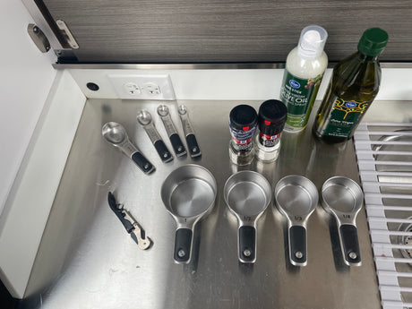 RV rental kitchen essentials: Measuring cups/spoons, oils, and spices on a compact stainless steel counter.