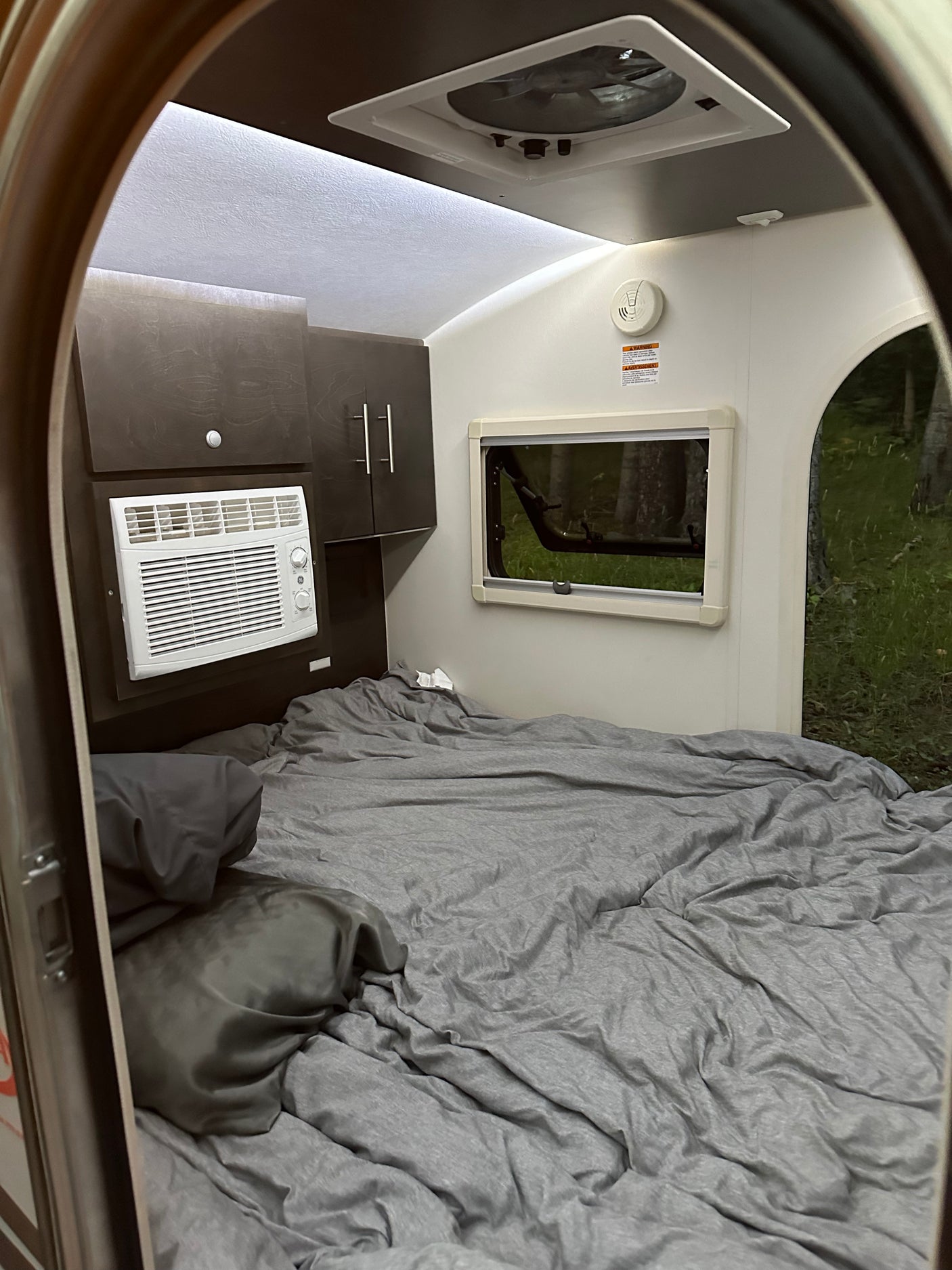 RV rental interior: a cozy bed with grey bedding, AC unit, and window showing trees outside.