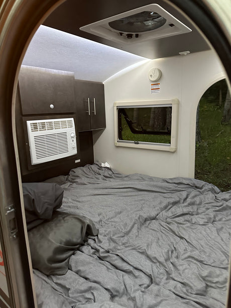 RV rental interior: a cozy bed with grey bedding, AC unit, and window showing trees outside.