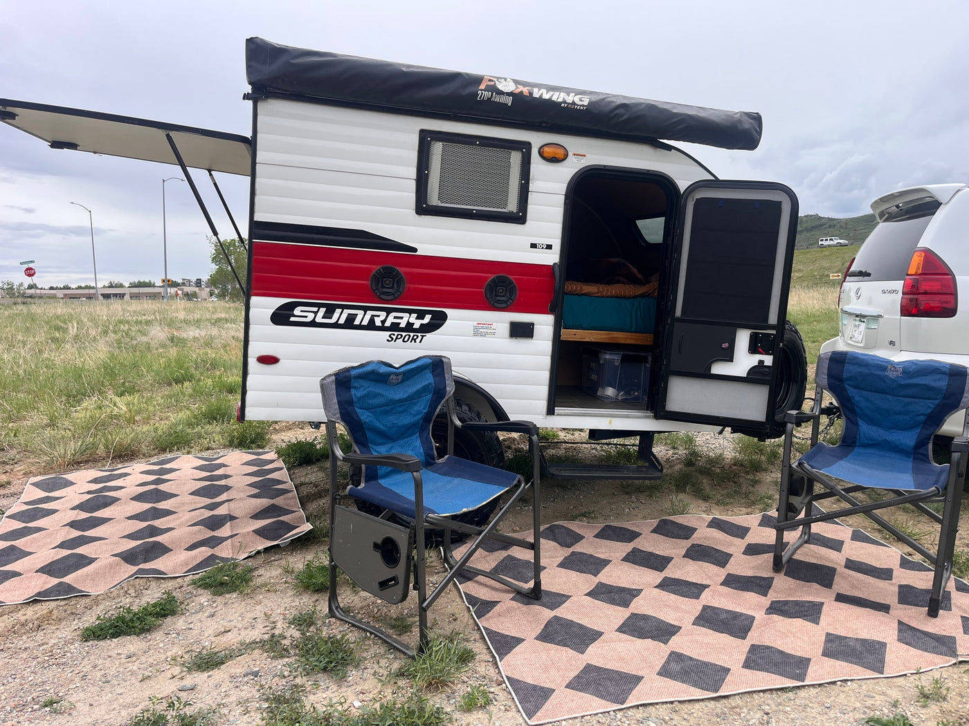 RV rental: A compact teardrop camper with an extended awning, camping chairs, and patterned mats awaits a relaxing stop. — photo 15