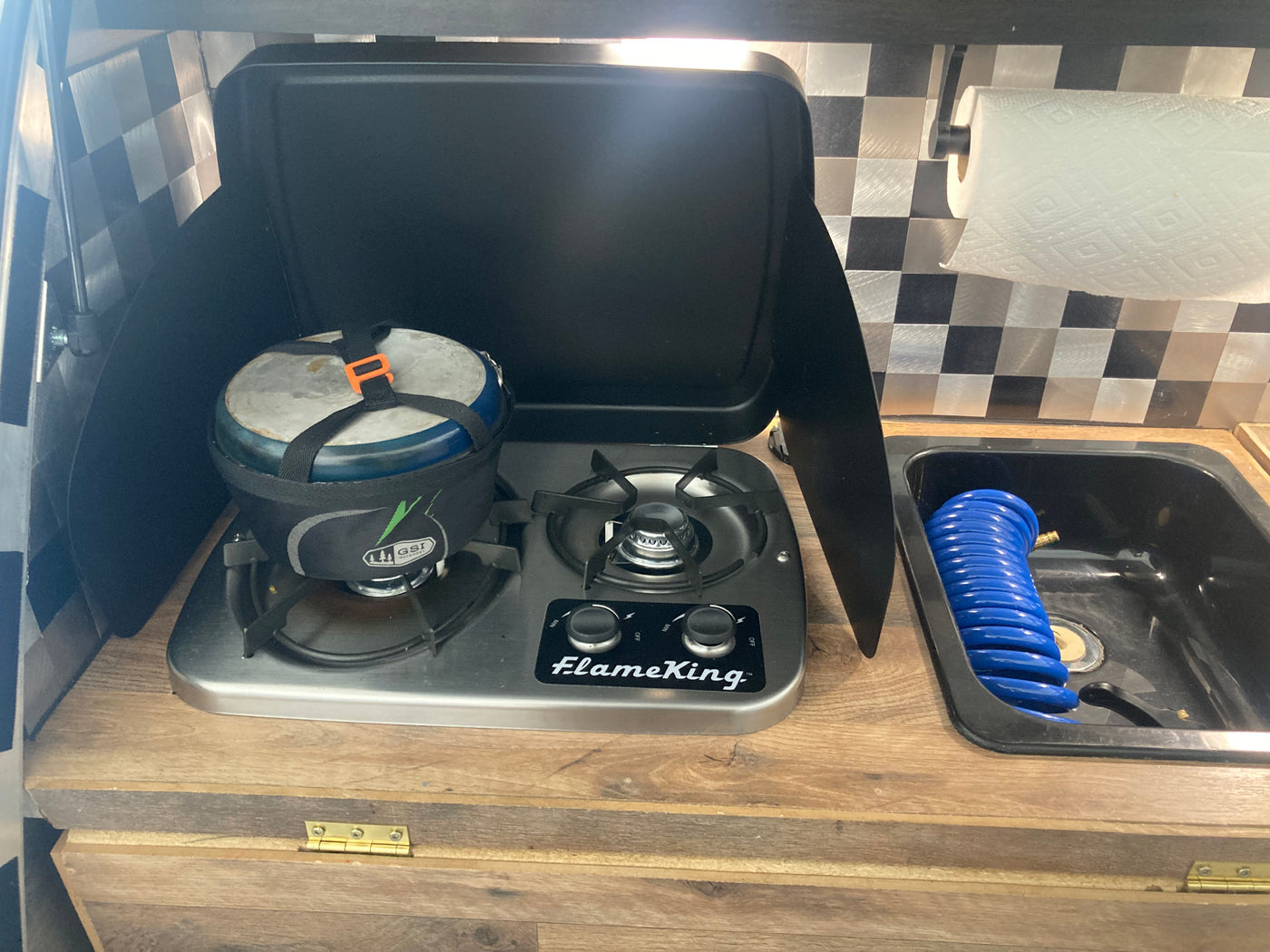 RV rental compact kitchen with a two-burner stove, a pot, sink with blue hose, and paper towel holder on a checkered wall. — photo 9