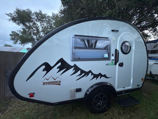Light gray nüCamp T@B Boondock teardrop rv rental, featuring mountain decals, parked on grass. Ready for adventure!